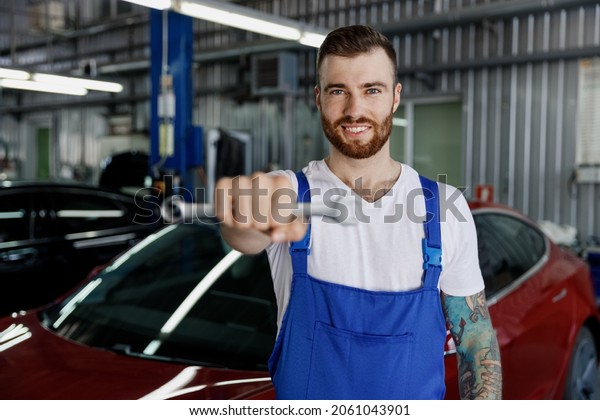 Smiling happy young fun professional technician\
car mechanic repairman man 20s in denim blue overalls white t-shirt\
hold give wrench key tool work in light modern vehicle repair shop\
workshop indoors