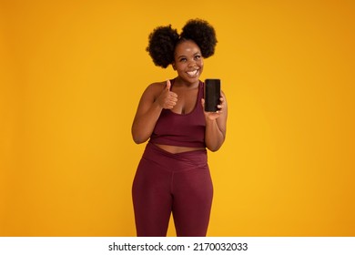 Smiling, Happy, Young Girl With Afro Hairstyle Posing On Yellow Studio Background In Sporty Clothes, Holding Mobile Phone In One Hand Showing New Fitness App. Okay Sign. Sale Concept. Copy Space. 