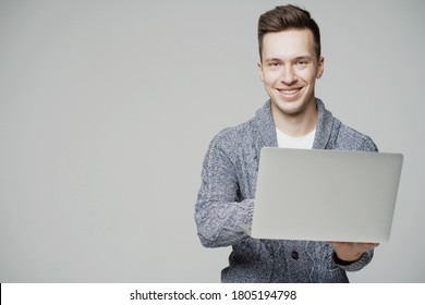 Smiling Happy Happy Young Guy Of European Appearance, Working On A Laptop. Grey Jacket, Good Mood. Solid Color Background