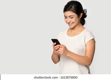 Smiling happy young indian ethnicity lady standing on right, aside blank empty free copy space for advertising text, looking at mobile phone screen, receiving good offer isolated on grey background.