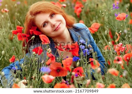 Smiling and happy young red-haired  girl in a blue denim jacket  enjoys the beauty and aroma of poppies in the field. Flower growing, floristics. Summer concept. Copy space