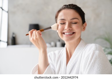 Smiling happy young woman applying makeup on her face with cosmetics brush in white spa bathrobe making morning preparations. Beauty care and treatment