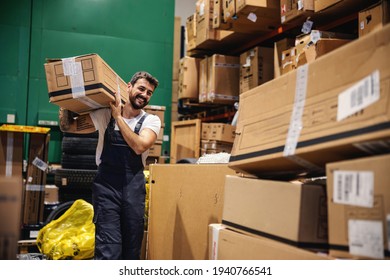 Smiling Hardworking Tattooed Bearded Blue Collar Worker In Overalls Carrying Box On His Shoulder And Preparing It For Export While Walking Trough Warehouse.