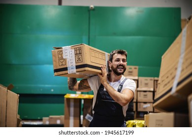 Smiling Hardworking Tattooed Bearded Blue Collar Worker In Overalls Carrying Box On His Shoulder And Preparing It For Export While Walking Trough Warehouse.