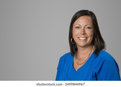 Smiling headshot of Caucasian brunette middle age medical worker woman in a blue scrubs top in a studio on gray grey backdrop - Powered by Shutterstock