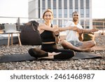 Smiling healthy athletic man and woman sitting on mats with legs crossed and stretching arms during morning exercise. Young couple doing sports on roof of modern building in morning.