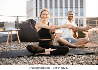 Smiling healthy athletic man and woman sitting on mats with legs crossed and stretching arms during morning exercise. Young couple doing sports on roof of modern building in morning. - Powered by Shutterstock