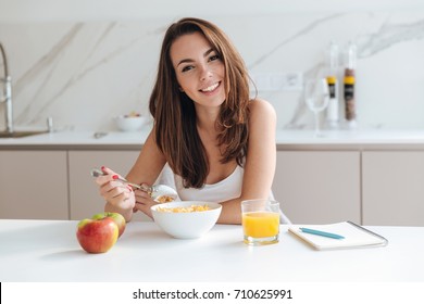 Smiling healthy woman eating corn flakes cereal while sitting and having breakfast at the kitchen table - Powered by Shutterstock