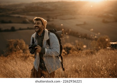 Smiling hiker tourist in the mountains at sunset. Man takes pictures of the sunset. Hipster with a photo camera and a backpack travels and enjoys nature. Copy space