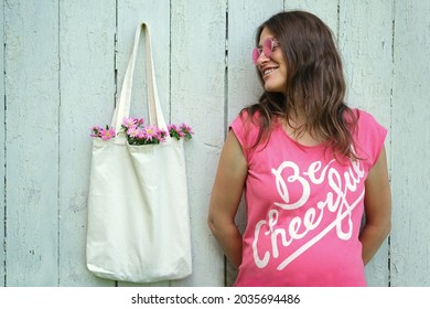 Smiling Hippie Woman In Pink T-shirt With Be Cheerful Inscription And Pink Round Glasses Standing Near Blank  Canvas Eco Shopping Bag With Flowers Inside. Zero Waste Mock-up.