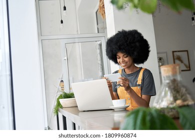 Smiling Hipster African American Female Student Using Laptop, Holding Phone Watching Mobile Streaming Ad, Tv Webinar, Remote Learning, Video Calling On Smartphone Sitting Alone At Table In Cozy Cafe.