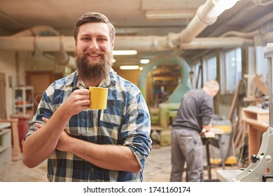 Smiling hipster carpenter with beard takes coffee break in his joinery