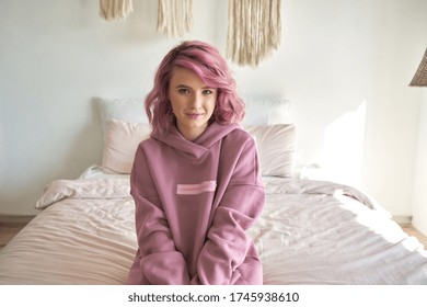 Smiling hipster gen z teen girl with pink hair and piercing wear hoodie looking at camera sitting on bed. Young millennial woman, adolescent teenager posing in cozy modern bedroom interior. Portrait