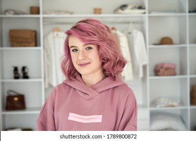 Smiling Hipster Gen Z Teen Girl Fashion Designer, Stylist With Pink Hair Piercing Wearing Hoodie Looking At Camera In Front Of Modern Clothes Wardrobe Closet, Face Head Shot Close Up Portrait.