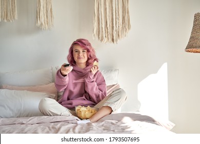 Smiling Hipster Gen Z Teen Girl With Pink Hair Watching Tv Movie Series Sitting In Bed, Holding Remote Control, Eating Chips Snack. Teenager Relaxing Watching Tv Television Reality Show In Bedroom.