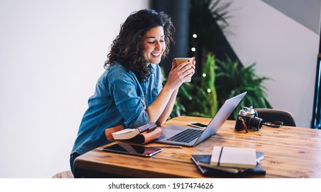 Smiling hipster girl in casual clothing watching positive video on website using laptop computer connected to wifi, cheerful female freelancer enjoying caffeine beverage while working remotely