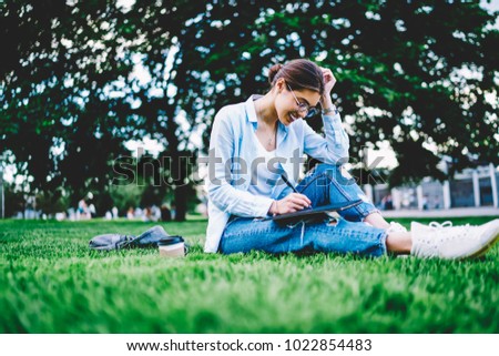 Smiling hipster girl in casual wear enjoying learning on green lawn in park writing report,positive young woman sitting on grass making creative drawings for project planning satisfied with spring day