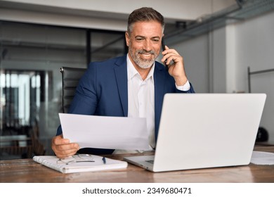 Smiling Hispanic businessman talking phone and check database in office. Happy Latin or Indian male business man holding documents, working at laptop computer doing online trade market tech research. - Powered by Shutterstock