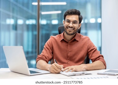 Smiling hispanic businessman working in modern office, confidently handling documents at desk. Demonstrates professional success and productivity with laptop - Powered by Shutterstock