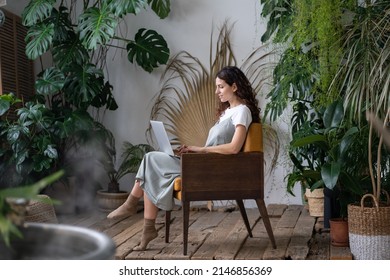 Smiling Hispanic Female Entrepreneur Sitting In Armchair In Greenhouse, Working On Laptop Surrounded By Exotic Houseplants. Pensive Italian Woman Typing On Computer. Cozy Workplace, Freelance Concept