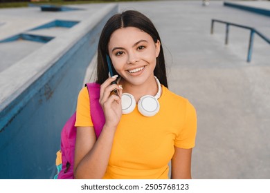 Smiling hispanic latin young teen girl school pupil student in casual with bag standing against ramp in city skate green park talking on mobile cell smart phone spending free time after school lessons - Powered by Shutterstock