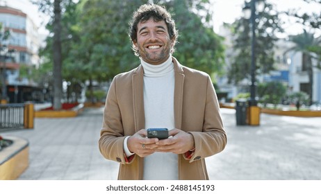 Smiling hispanic man with beard using smartphone in sunlit city park. - Powered by Shutterstock