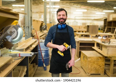 Smiling Hispanic Man Doing Carpentry Work In A Woodshop. Happy Carpenter Working With A Power Drill And A Hammer