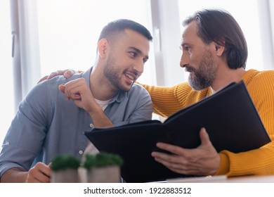 Smiling Hispanic Man Looking At Father Holding Photo Album On Blurred Foreground