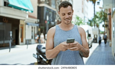Smiling hispanic man with tattoos using smartphone on sunny urban street.