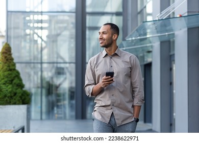 Smiling hispanic man walking down street near modern office building, freelancer businessman looking away holding mobile phone. - Powered by Shutterstock