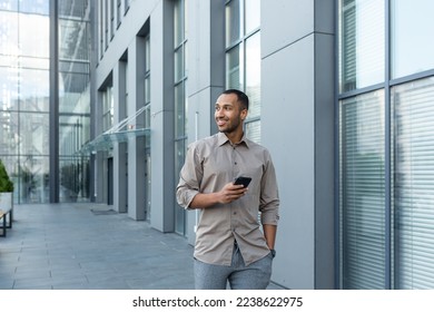 Smiling hispanic man walking down street near modern office building, freelancer businessman looking away holding mobile phone. - Powered by Shutterstock