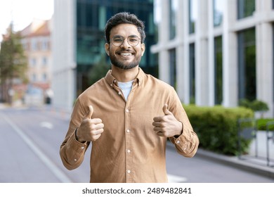 Smiling hispanic man wearing casual outfit giving thumbs up on city street. Young professional showing positivity, confidence, and approval outdoors urban environment. Modern buildings background. - Powered by Shutterstock