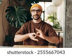 Smiling Hispanic man wearing yellow hat and eyeglasses showing sign language gesture looking at camera. Communication concept