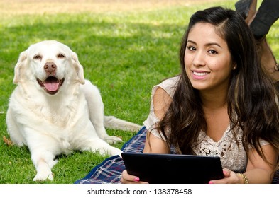 A Smiling Hispanic Woman In The Park With Her Pet Dog And Holding A Tablet.