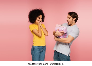 Smiling Hispanic Woman Touching Face While Looking At Amazed Man With Plenty Of Presents Isolated On Pink