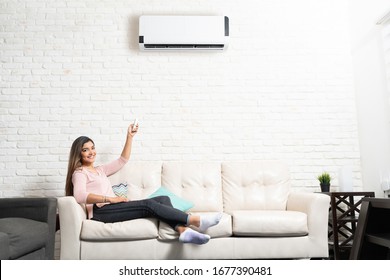 Smiling Hispanic young woman adjusting temperature of air conditioner while sitting on sofa at home - Shutterstock ID 1677390481