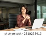 Smiling Hispanic young woman entrepreneur, happy female executive manager looking at camera sitting at workplace with laptop. Portrait of confident businesswoman leader in her 30s at work desk.