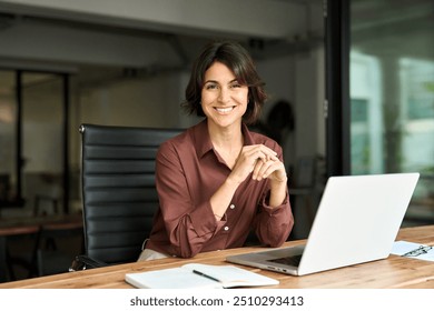 Smiling Hispanic young woman entrepreneur, happy female executive manager looking at camera sitting at workplace with laptop. Portrait of confident businesswoman leader in her 30s at work desk.