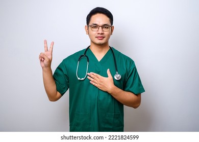 Smiling Honest Asian Male Nurse Or Doctor, Intern In Scrubs Giving Pledge, Raising One Arm And Hold Hand On Heart While Promise, Oath To Patient Over White Background