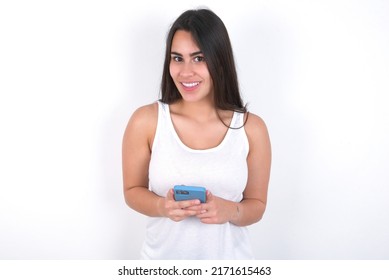 Smiling Honest Young Beautiful Brunette Woman Wearing White Top Over White Wall Friendly And Happily Holding Mobile Phone Taking Selfie In Mirror.