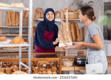 Smiling hospitable Muslim woman, bakery owner, dressed in blue khimar and apron, engaging in friendly conversation over counter and offering fresh baguettes to young female customer - Powered by Shutterstock