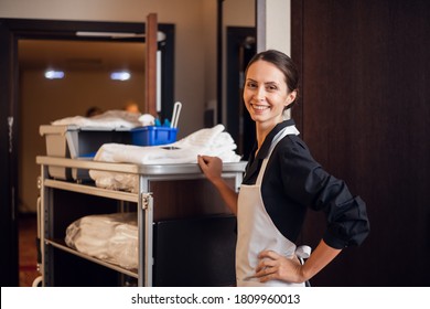 A smiling hotel maid with cleaning cart and cleaning supplies, looking at the camera and posing. - Shutterstock ID 1809960013