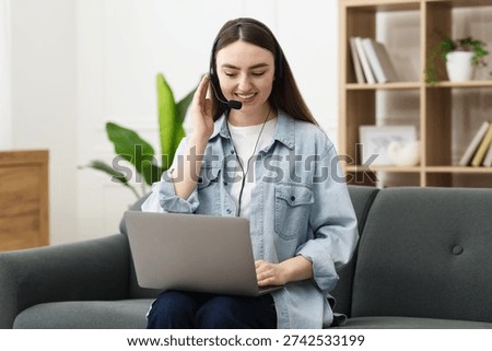 Smiling hotline operator in headset having videocall with client by laptop on sofa indoors