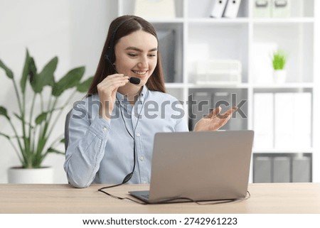 Smiling hotline operator in headset having videocall with client by laptop at table in office