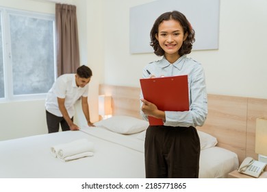 Smiling Household Manager With Clipboard Note On Background Of Maid Cleaning Hotel Room