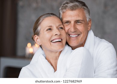 Smiling husband embracing cheerful wife from behind at spa. Laughing mature couple enjoying a romantic hug at wellness center after massage. Senior man and woman in white in bathrobe relaxing at spa.