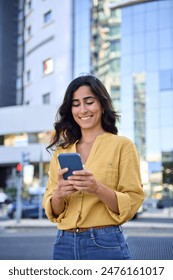 Smiling indian arabic student woman in casual shirt holding cellphone for online business work application. Young middle eastern Israel businesswoman using smartphone mobile phone outdoors. Vertical 