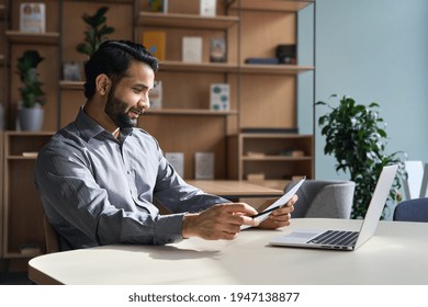 Smiling indian business man employee working on laptop looking at document at home office. Male ethnic professional teacher reading paper, employer holding cv, lawyer checking contract sits at table.