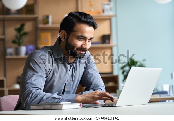 Smiling indian business man working on laptop at\
home office. Young indian student or remote teacher using computer\
remote studying, virtual training, watching online education\
webinar at home office.