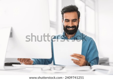 Smiling indian businessman in blue shirt reading paper with satisfaction while working at his computer station in well-lit office interior